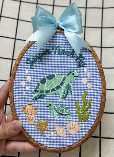Real embroidered ornament with a blue gingham background, light blue bow and a stitched name